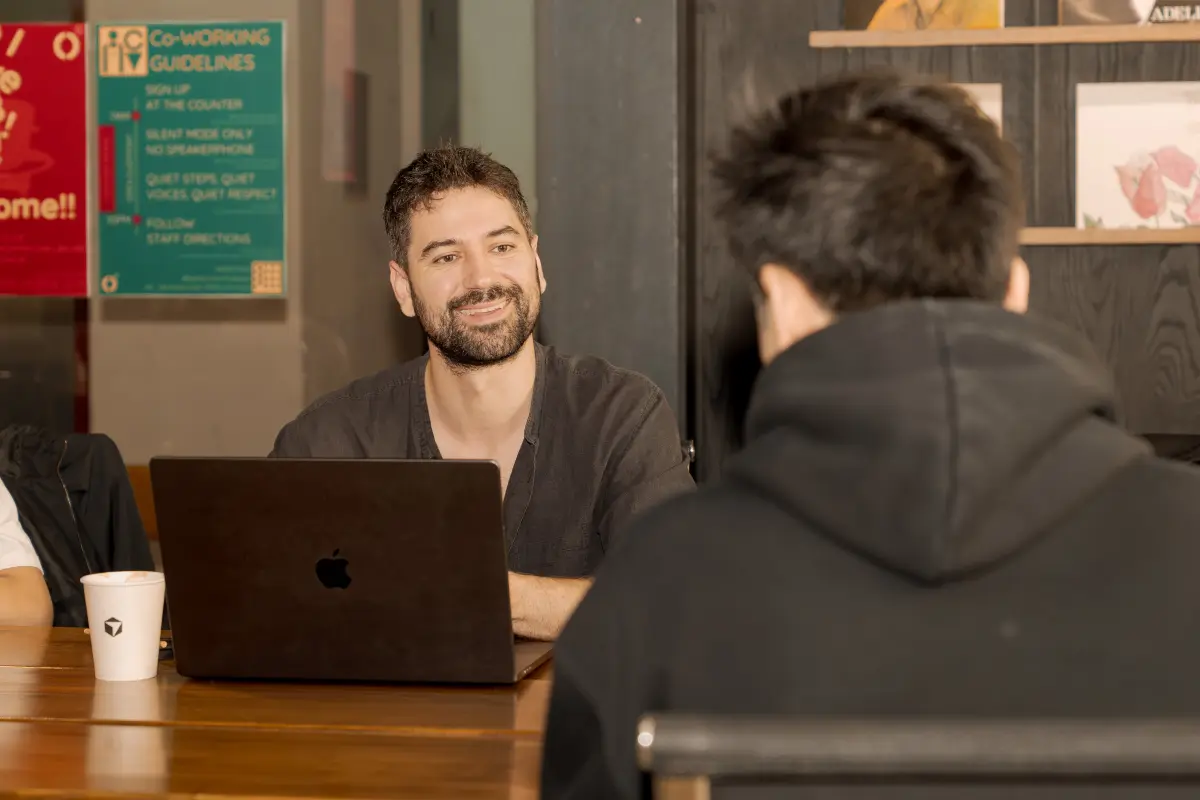 A bearded builder smiling mid-conversation at a laptop during a Cursor Da Nang coworking meetup